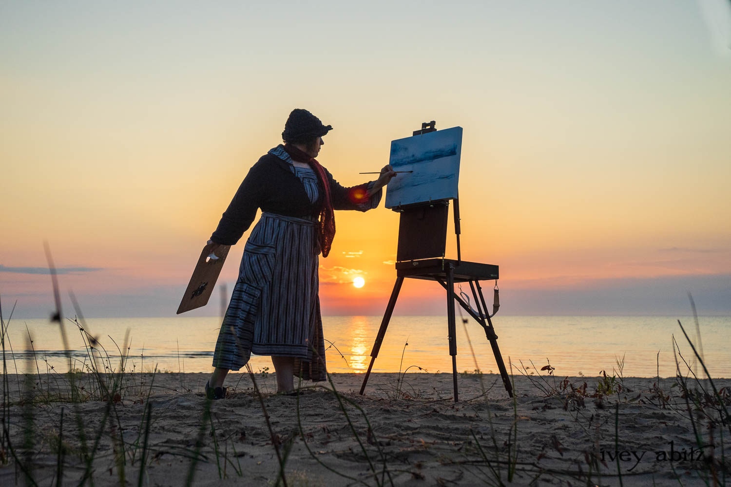 Cynthia Ivey Abitz painting the Lake Huron sunrise. Beacon Jacket in Majestic Blue Stripe Knit Beacon Dress in Majestic Blue Variegated Stripe Weave Cilla Slip Frock in Signature Black Bamboo Silky Knit Hapgood Hat in Majestic Blue and Mountain Checked Hat Weave Baedeker Scarf in Crimson Sky Mohair Hat and Scarf Knit Ivey Abitz Dog Tag Necklace
