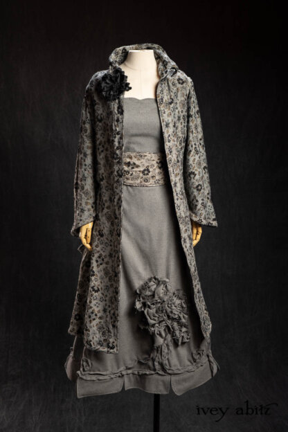 Limited Edition Phinneus Coat in Black Diamond and Pewter Floral Brocade; Limited Edition Porte Cochere Sash in Black Diamond and Pewter Floral Brocade; Chevallier Frock in Pewter Cashmere Cotton Twill; Bonheur Brooch in Black Sculpted Felt; Cilla Slip Frock in Signature Black Washed Silk Jersey Knit. - Bespoke clothing by Ivey Abitz.